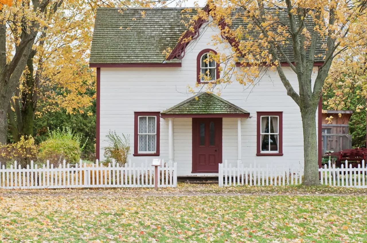 A white house with a small front porch, a white picket fence, and autumn trees